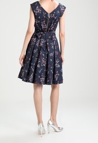 Marinblommig klänning med korta ärmar, V-ringning bak och knytning i midjan. Kjolen vidgar sig, tyget har en slät textur. Silverfärgade högklackade skor.