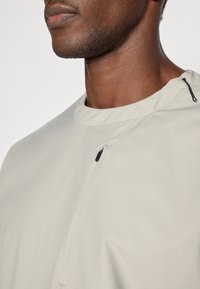 Haut gris clair avec une coupe décontractée, un col rond et un détail de fermeture éclair noire au niveau du col. Texture lisse avec des coutures minimales visibles.