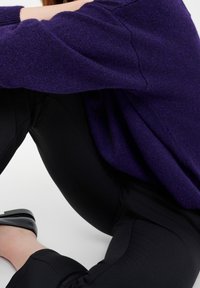 Personne assise, jambes pliées, portant un pantalon noir, des chaussures noires et un pull violet, sur un fond clair uni.