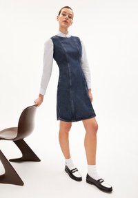 Robe en denim à la silhouette ajustée, de couleur bleue, avec des coutures visibles. Portée avec une chemise blanche, des mocassins noirs et des chaussettes à la cheville. Chaise marron en arrière-plan.