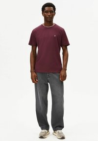 Bordeauxrode T-shirt met korte mouwen, een geribde halslijn en een klein logo op de borst, gecombineerd met loszittende grijze jeans en sneakers.