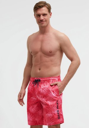 Oberkörperfreier Mann trägt rote Badeshorts mit tropischem Muster, schwarzem Kordelzug und der Aufschrift "Camp David" am linken Bein, steht mit der Hand in der Tasche.