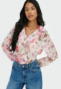 Blusa de gasa floral con escote con volantes, acentos en rosa y mangas largas, combinada con jeans de talle alto en azul. El fondo es blanco liso.