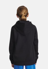 Personne aux cheveux blonds en queue de cheval portant un sweat noir et un pantalon bleu, tourné le dos contre un fond blanc uni.