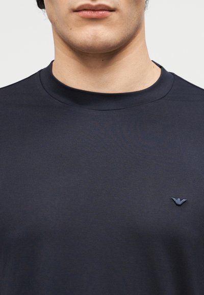 Άνδρας που φοράει μπλούζα σκούρου ναυτικού με στρογγυλή λαιμόκοψη και μικρό κεντημένο έμβλημα στο αριστερό στήθος, εμφανίζοντας το κάτω μέρος του προσώπου και τον λαιμό.