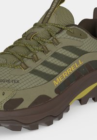 Πράσινο και καφέ παπούτσι Merrell trail με κίτρινο λογότυπο, ετικέτα Gore-Tex, πάνω μέρος από διχτυωτό υλικό και ανθεκτική σόλα για πεζοπορία σε εξωτερικούς χώρους.