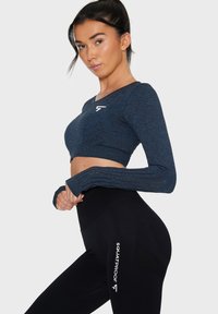 Langärmliges, dunkelblaues Crop-Top mit strukturierter Oberfläche und Daumenschlaufenärmeln, kombiniert mit hochgeschnittenen schwarzen Leggings, die seitlich mit "SQUATPROOF" bedruckt sind.