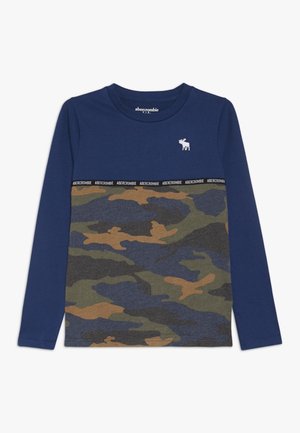 Chemise à manches longues avec une partie supérieure bleue et une partie inférieure à motif camouflage en verts, bruns et bleus, avec un petit logo.