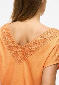 Pomarańczowa bluzka z dekoltem w kształcie litery V, ozdobiona skomplikowanym okrągłym haftem oraz teksturowanym materiałem. Krótkie rękawy i luźny krój.