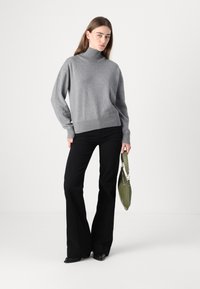 Un pull à col roulé gris en tricot doux, associé à un pantalon noir évasé. La modèle tient un sac vert, se tenant devant un fond blanc uni.