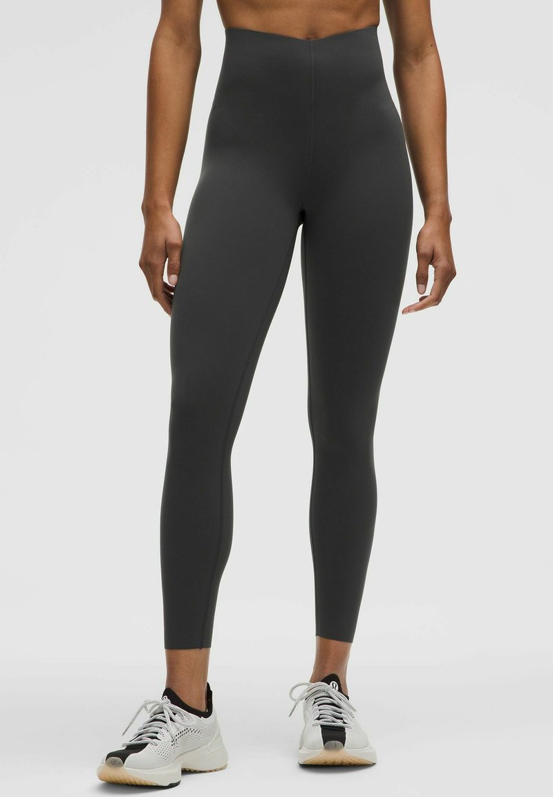Legginsy o wysokim stanie, dopasowane, w ciemnoszarym kolorze, wykonane z elastycznego materiału o gładkiej fakturze. Połączone z białymi butami sportowymi.