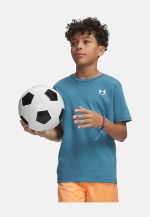 Kihara juustega poiss kannab türkiissinist Under Armour T-särki ja oranžeid pükse, hoides valgel taustal mustvalget jalgpalli.