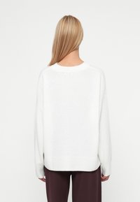 Balts, mīksts, pārāk liels džemperis ar apaļu kakla izgriezumu un ribotu apakšmalu, ar garām piedurknēm un brīvā stila piegriezumu, apvienots ar tumšām biksēm.