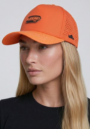 Woman with long blonde hair wearing a bright orange baseball cap with "BLACKSKIES PRO" logo and a black shirt, looking slightly left.