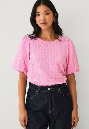 Kvinde iført en tekstureret, lyserød bluse med korte ærmer og mørke, højtaljede jeans, stående foran en ensartet lys baggrund.