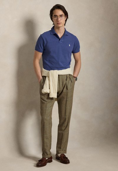 Ung mann med blå polotrøye, rutete bukser, brune loafers, kremfarget genser knyttet rundt livet, står foran en ensfarget bakgrunn med hendene i lommene.