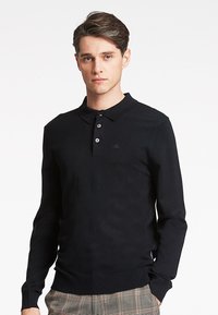Musta pitkähihainen polo-paita, jossa on kaulus, kolme nappia ja huomaamaton logo. Sileä kangas, jossa on ribbaillut hihansuut ja helma.
