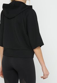 Černá zkrácená mikina s kapucí, širokými rukávy a strukturovanou tkaninou. Vhodná pro aktivní nošení, doplněná černými legínami s síťovinovými detaily.