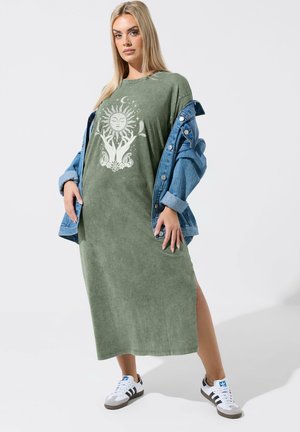 Donna che indossa un vestito midi verde con grafica di sole e luna, giacca di jeans scesa sulle spalle e sneakers bianche Adidas con strisce nere.