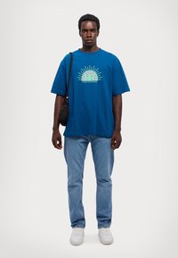 Blå bomull T-skjorte med korte ermer og grafisk trykk. Brukt med lyseblå jeans og hvite sneakers, samt en liten svart skulderveske.