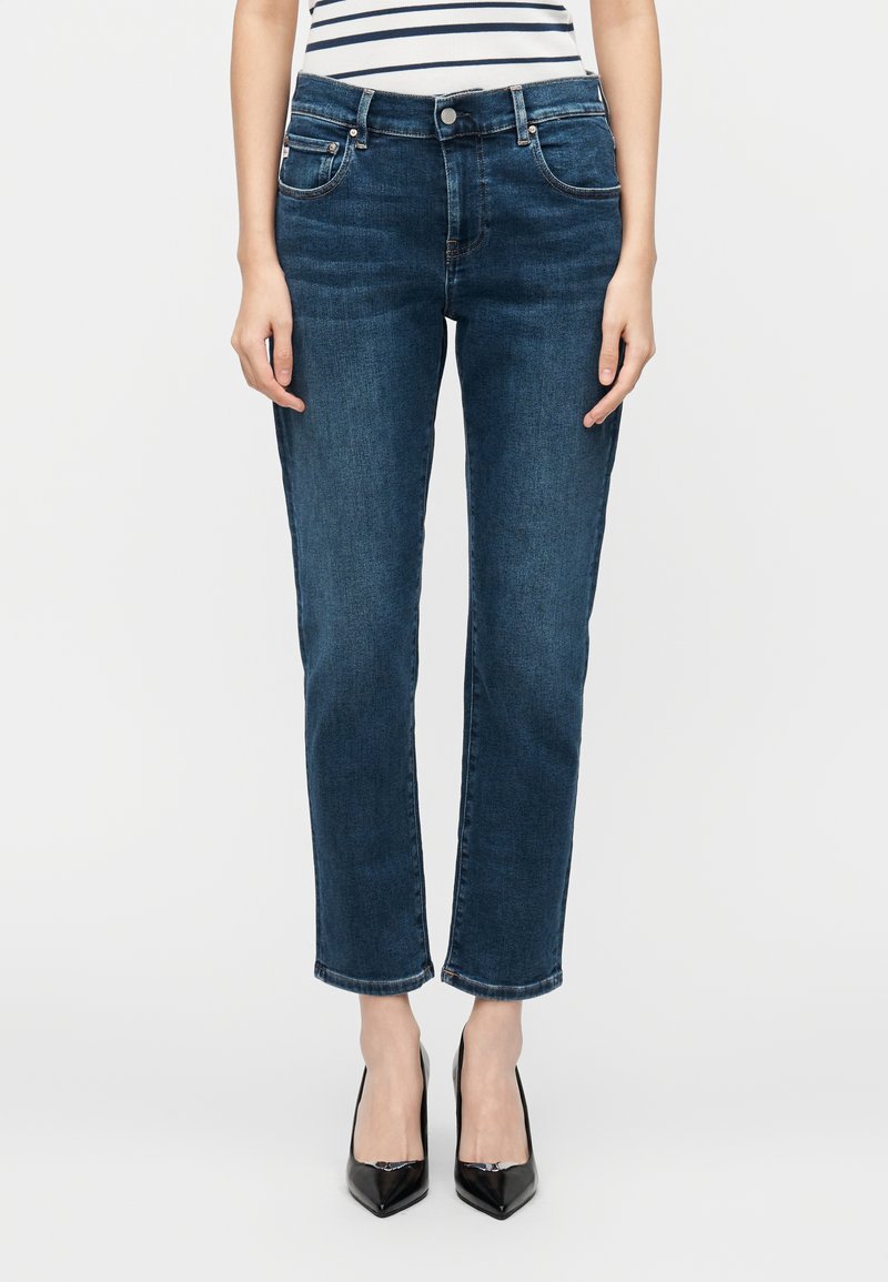 Tamnoplave traperice ravnog kroja s pet džepova i uvrnutim rubovima, uparene s crnim cipelama špicasto vrha.