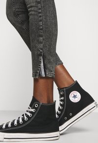Zapatillas altas de lona negras con cordones blancos y suelas de goma. Vaqueros ajustados grises con una abertura lateral y una etiqueta visible en el dobladillo.