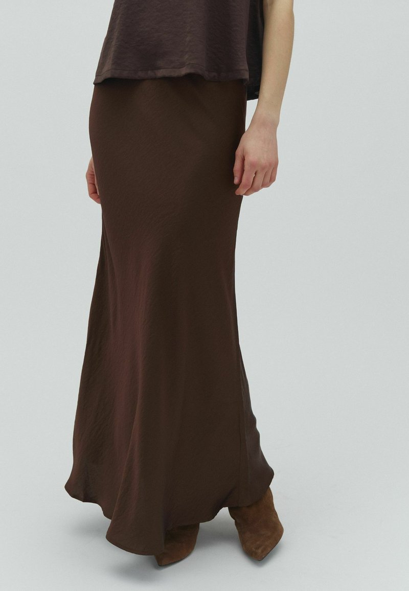 Femme portant une jupe longue fluide marron foncé avec un haut assorti et des bottes pointues en daim marron, debout devant un fond uni.