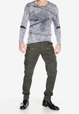 Uomo che indossa una maglia grigia a maniche lunghe con dettagli di zip neri, pantaloni cargo verde oliva con zip e stivali neri lucidi.