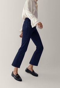 Mörkblå cropped jeans med rak benpassform, i kombination med en vit blus med trumpetärmar och svarta loafers med gulddetaljer.