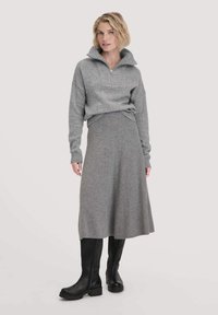 Frau in einem grauen Reißverschluss-Sweater, grauem Midirock und schwarzen kniehohen Stiefeln, die vor einem einfachen hellen Hintergrund steht.