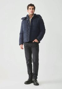 Man wearing navy padded jacket, black pants, and black boots standing against a plain light background.