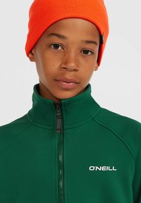 Groene ritssluiting jack met hoge kraag en O'Neill logo, gecombineerd met een feloranje gebreide beanie. Zachte textuur, aansluitend ontwerp.