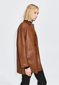Bruine leren jas met een kraag, voorzien van twee zwarte knopen, een zijzak en een relaxed fit. Glad textuur en een middellange pasvorm.