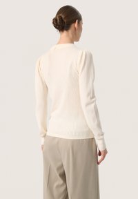 Ribbed cream sweater with puffed long sleeves, featuring a rounded neck and fitted cuffs, worn with beige wide-leg trousers.