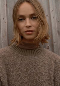 Strukturierter, brauner Strickpullover mit einem gerippten Kragen, über einem braunen Oberteil getragen, vor einem rustikalen Holz-Hintergrund. Subtile Nuancen in den Farbtönen.