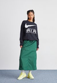 Sweatshirt noir Nike avec un grand logo blanc, associé à une jupe verte dotée d'un cordon de serrage ajustable. Des chaussures de sport jaunes viennent compléter la tenue.