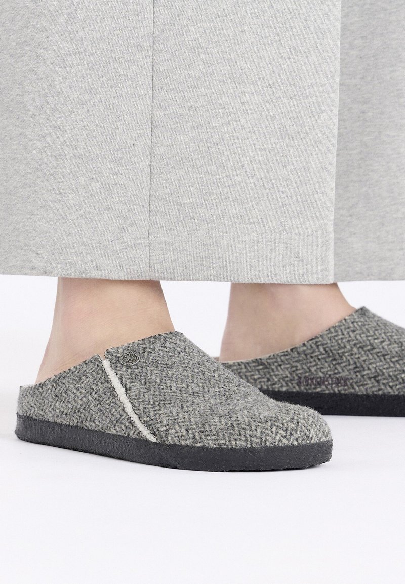 Chaussons en laine grise avec un motif en chevrons, dessus en tissu doux, semelle noire contrastante et détails de couture subtils.