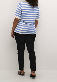 Gestreiftes Strickoberteil in Blau und Weiß mit kurzen Ärmeln, kombiniert mit schwarzen, eng anliegenden Hosen und schwarzen Sandalen mit Absatz. Schlicher Hintergrund.