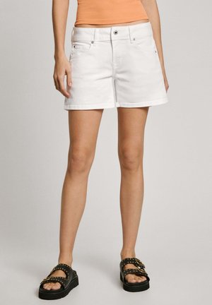 Person iført hvide denimshorts, orange ærmeløs top og sorte sandaler med gyldne spænder mod en ensfarvet baggrund.