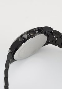 Montre en acier inoxydable noir avec une finition lisse. Elle présente deux poussoirs et une couronne texturée, un bracelet à maillons et un fond argenté poli.