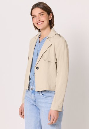 Femme souriante portant une veste courte beige sur une chemise en denim bleu clair et un jean, debout devant un fond clair uni.