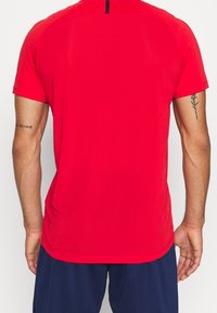 Hombre que lleva una camisa de mangas cortas roja brillante y pantalones azul marino, visto desde atrás con tatuajes en ambos brazos superiores.