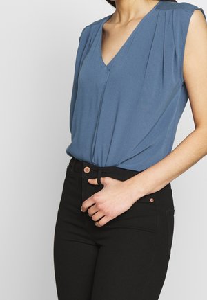 Frau trägt eine ärmellose blaue V-Ausschnitt-Bluse, die in eine hoch taillierte schwarze Hose mit kupferfarbenen Knopfdetails gesteckt ist.