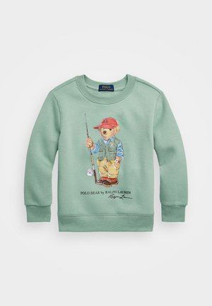 Sudadera verde menta con una ilustración de un oso que sostiene una caña de pescar, con una gorra roja, chaleco y pantalones caqui. En la parte inferior incluye texto con la marca.
