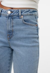 Šviesiai mėlynos džinsinės kelnės su vidutinio aukščio juosmeniu, priekinėmis kišenėmis ir vario spalvos detalėmis. Audinys rodo nežymų nudilimą ir yra lygus.