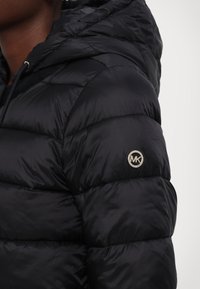 Chaqueta de plumas negra con capucha, diseño acolchado, textura brillante y un logo plateado de MK en el brazo izquierdo. Cierre con cremallera y cordón ajustable.