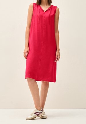 Czerwona sukienka bez rękawów, do kolan, z dekoltem w serek i sznurkiem do regulacji. Miękka, teksturowana tkanina; noszona z białymi i bordowymi sneakersami.