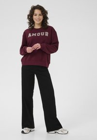 Felpa bordeaux con il testo "AMOUR" di colore chiaro, abbinata a pantaloni neri a gamba larga. Tessuto morbido, vestibilità rilassata e maniche a polsino.