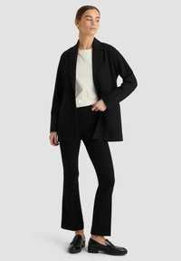 Schwarzer maßgeschneiderter Blazer über einem weißen Oberteil, kombiniert mit schwarzen ausgestellten Hosen und schwarzen Loafers. Glatter Stoff und strukturierte Designs.