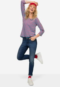 Pasiasty, długi rękaw w kolorze turkusowym i czerwonym, dżinsowe spodnie, białe trampki i czerwone skarpetki. Model pozuje z uniesioną jedną nogą i uniesioną ręką.